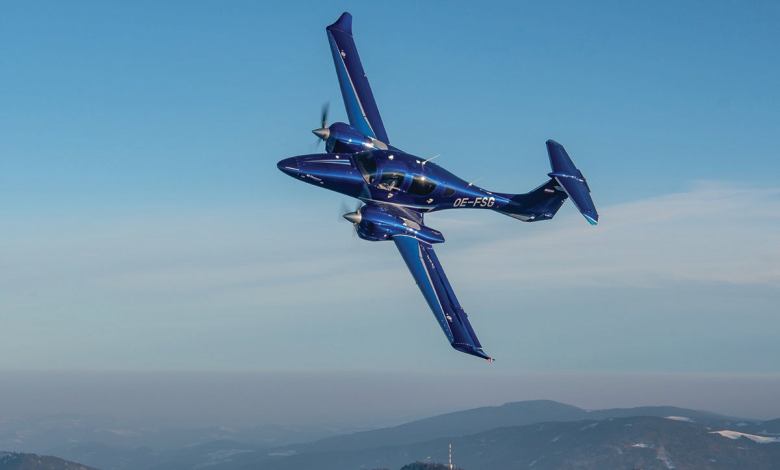 A Diamond D62 airplane soars through clear skies over low mountains in the background.