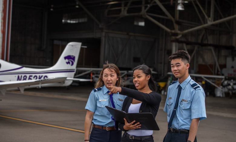 A Black female student points at the tarmac as two CFIs look on. A training aircraft and a hangar are in the background