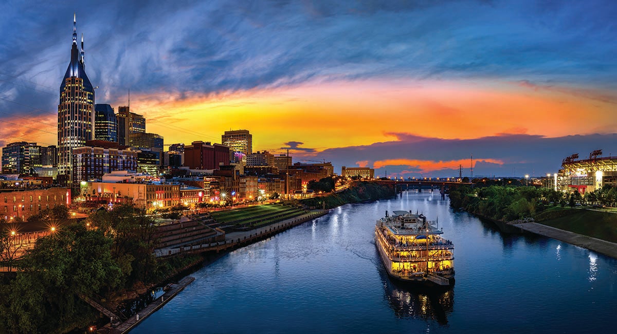 The city of Nashville at sunset. A paddle wheel cruise ship cruises down the Cumberland River amid a backdrop of the downtown area.