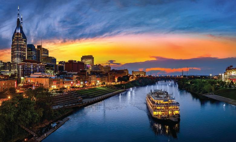 The city of Nashville at sunset. A paddle wheel cruise ship cruises down the Cumberland River amid a backdrop of the downtown area.