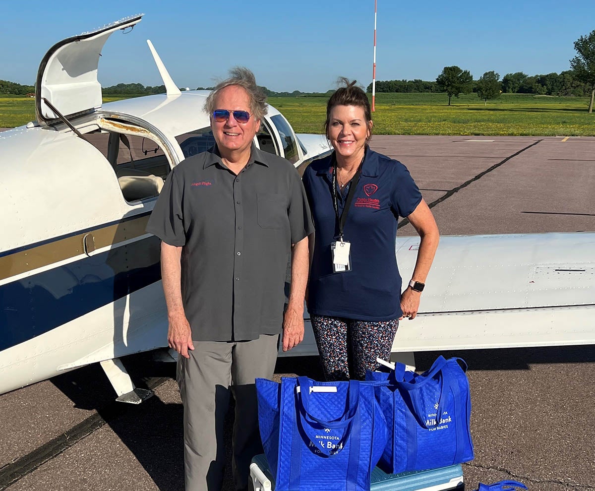 Two people stand in front of a Mooney aircraft on a tarmac with bags of breast milk to be delivered to medically fragile babies.
