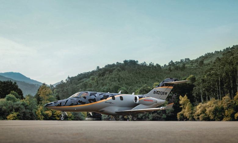 A HondaJet Elite S airplane in a gray camouflage livery sits on a tarmac with green trees and hills in the background
