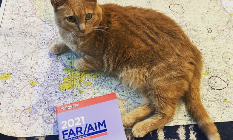 A ginger cat lies across an aeornautical sectional chart near a copy of the FAR/AIM