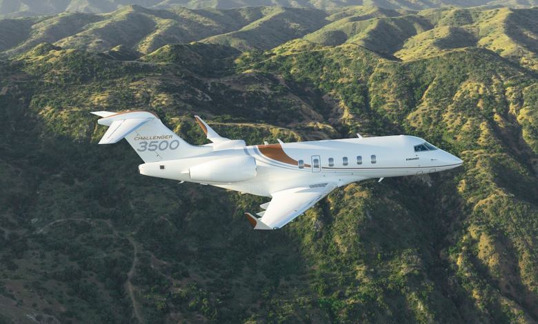 A Bombardier Challenger 3500 flies over a hilly, woodsy landscape.