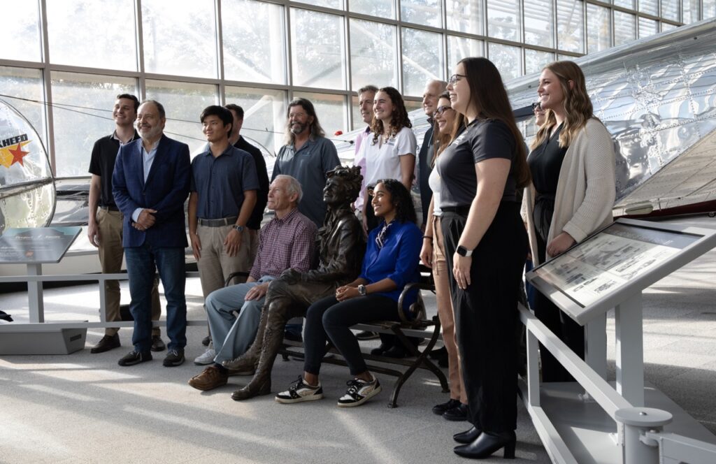 Fourteen of the 15-member expedition team poses for a photo, with team leader Richard Pettigrew seated in front on a bench next to a statue of Amelia Earhart.  [Courtesy: Ted Huetter/Museum of Flight]
